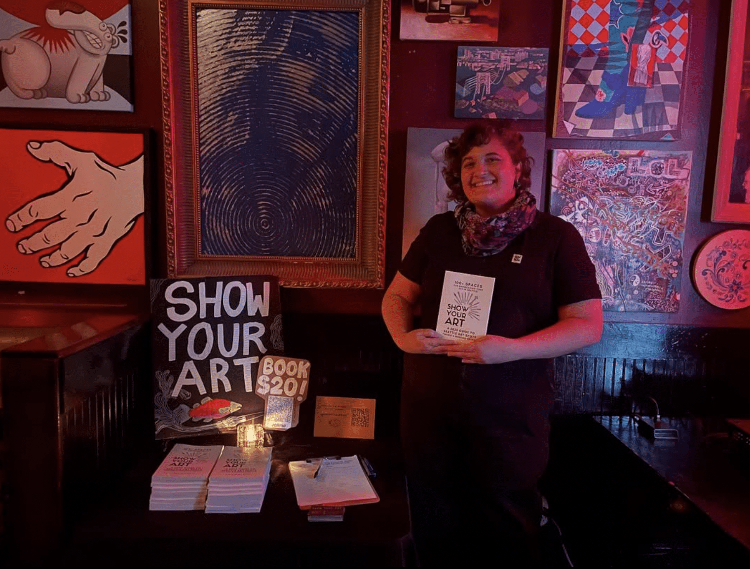 A person stands beside a table displaying books titled "Show Your Art" for $20, surrounded by colorful artwork on a dimly lit wall.