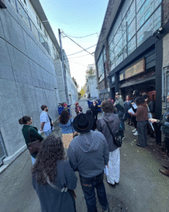 A group of people stand in an alleyway between industrial buildings, listening to a person speaking while holding papers.