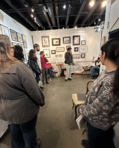 A group of people stands inside an art gallery, observing framed artworks displayed on the walls while a person seated to the right gestures during a discussion.