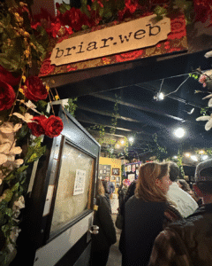 People stand in line inside a decorated space with flowers and vines, under a sign that reads "briar.web." A QR code is posted on the open door. The room is warmly lit.