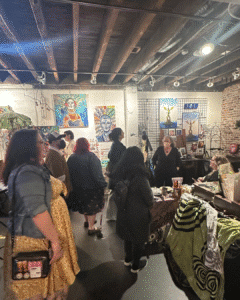 People browse art and merchandise at an indoor market with exposed beams, colorful paintings on the walls, and tables covered in items for sale.