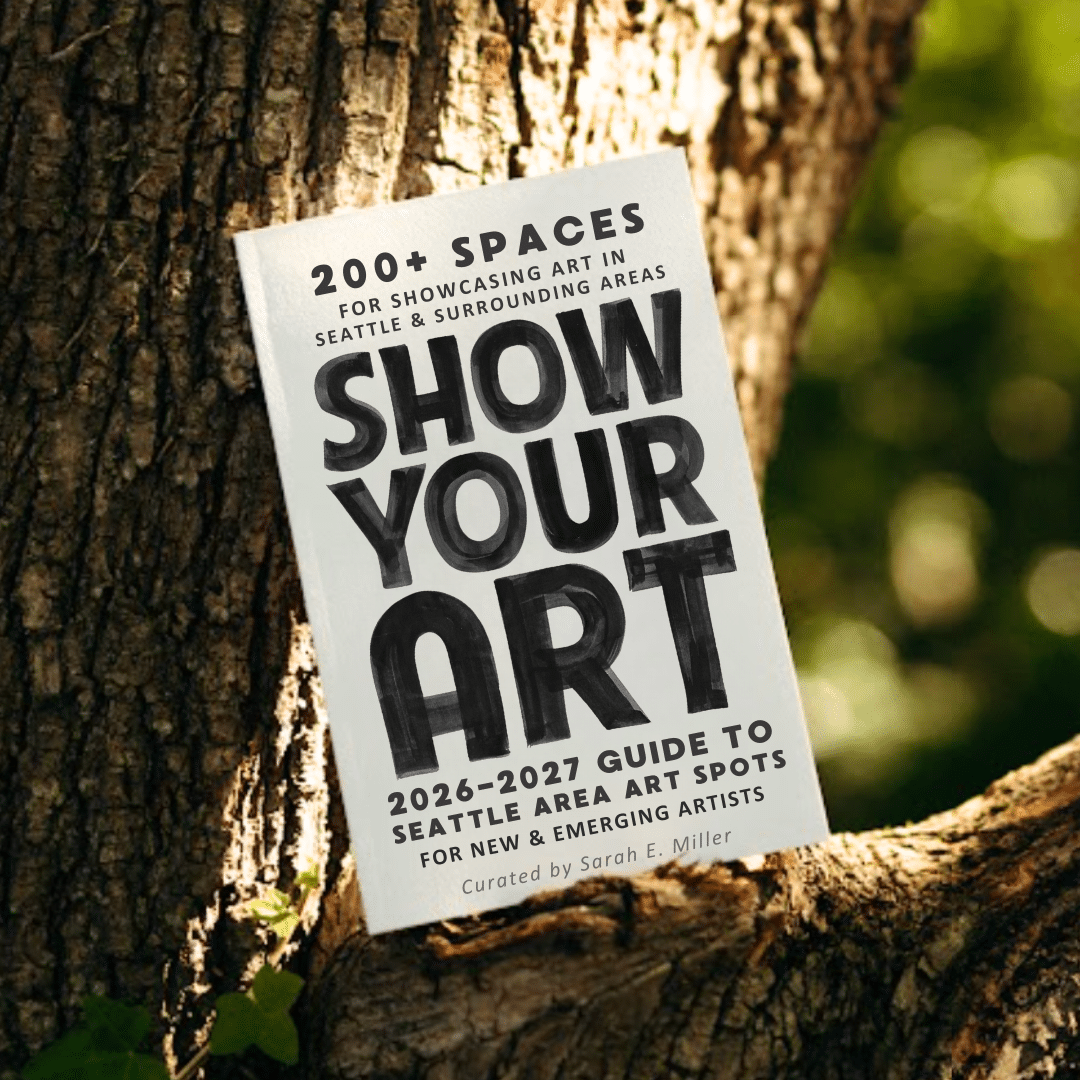 A book titled "Show Your Art: 2026-2027 Guide to Seattle Area Art Spots" leans against a tree trunk outdoors.