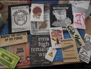 A tabletop displays various illustrated zines, stickers, and cards with different titles, artwork, and designs arranged on a cutting mat.