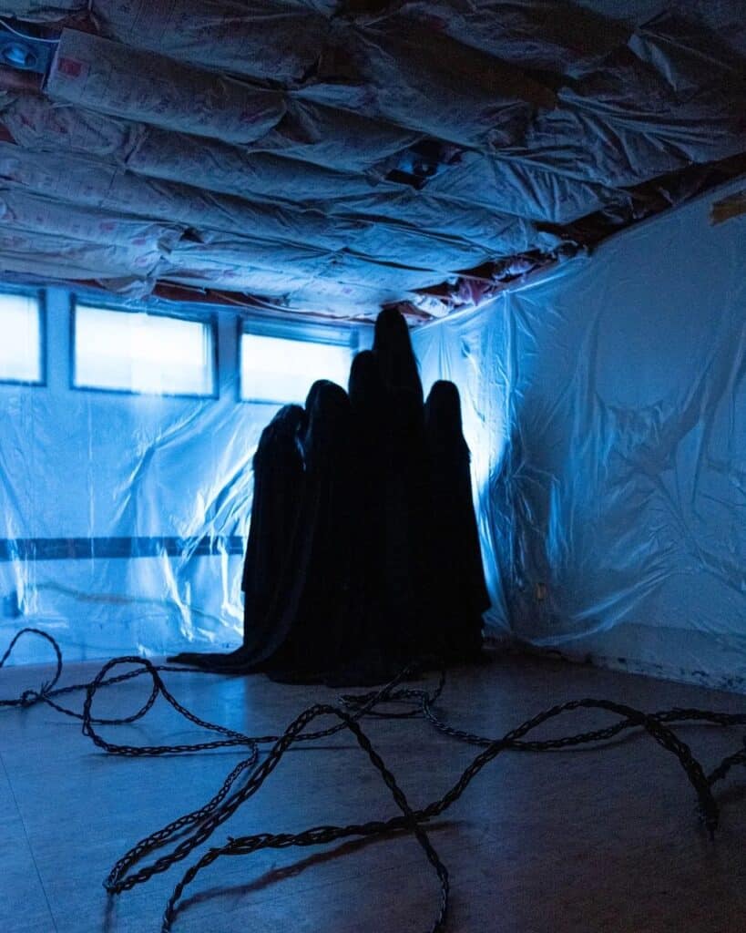 A group of figures in black cloaks stands together in a dimly lit room with plastic-covered walls, exposed insulation, and chains strewn across the floor.