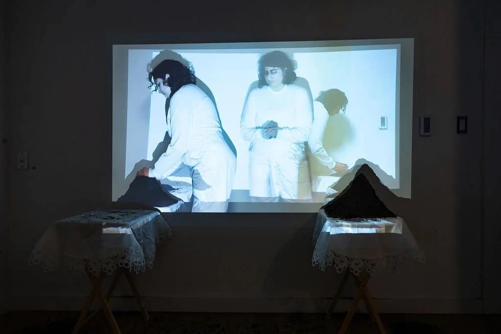 A video is projected onto a wall showing a person in white, while two small tables with lace cloths and dark objects are positioned beneath the projection.