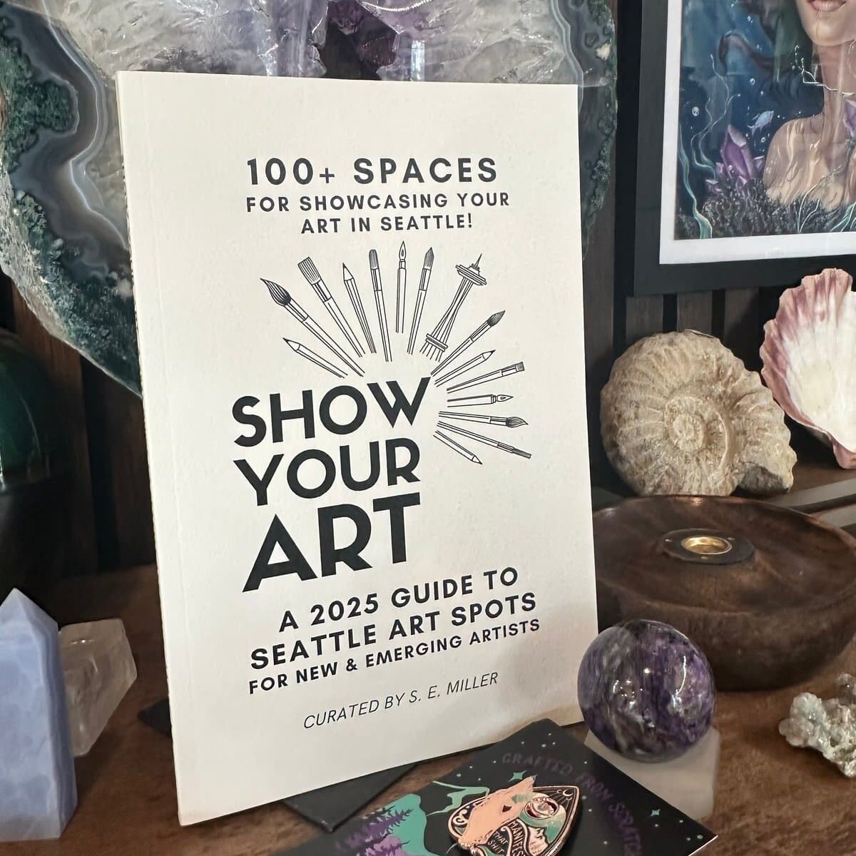 A book titled "Show Your Art: A 2025 Guide to Seattle Art Spots" stands on a wooden shelf surrounded by crystals, rocks, and artwork.