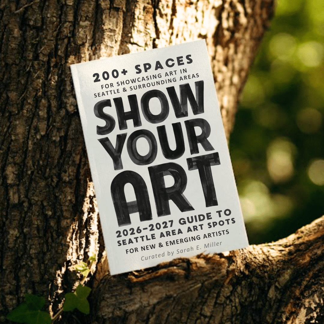 A book titled "Show Your Art: 2026-2027 Guide to Seattle Area Art Spots" rests against the trunk of a tree outdoors.